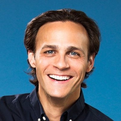 A headshot photograph of a smiling man with dark brown hair and blue eyes, wearing a dark blue button-down shirt against a solid blue background.