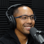 A close-up profile shot of a smiling man in a podcast studio, wearing glasses and black headphones. He is speaking directly into a Shure SM7B microphone, with a wall of black acoustic foam panels visible in the background.