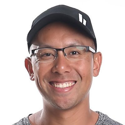 A headshot photograph of an East Asian man with a shaved head, wearing black-framed glasses and a black baseball cap with a white graphic logo on the side. He is smiling broadly, showing his teeth, and is wearing a grey heathered t-shirt. The background is plain white.