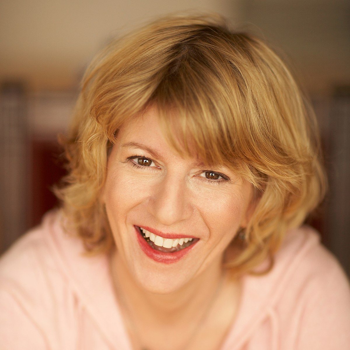 A headshot photograph of a smiling middle-aged woman with shoulder-length wavy blonde hair and brown eyes, wearing a pink hooded sweatshirt.
