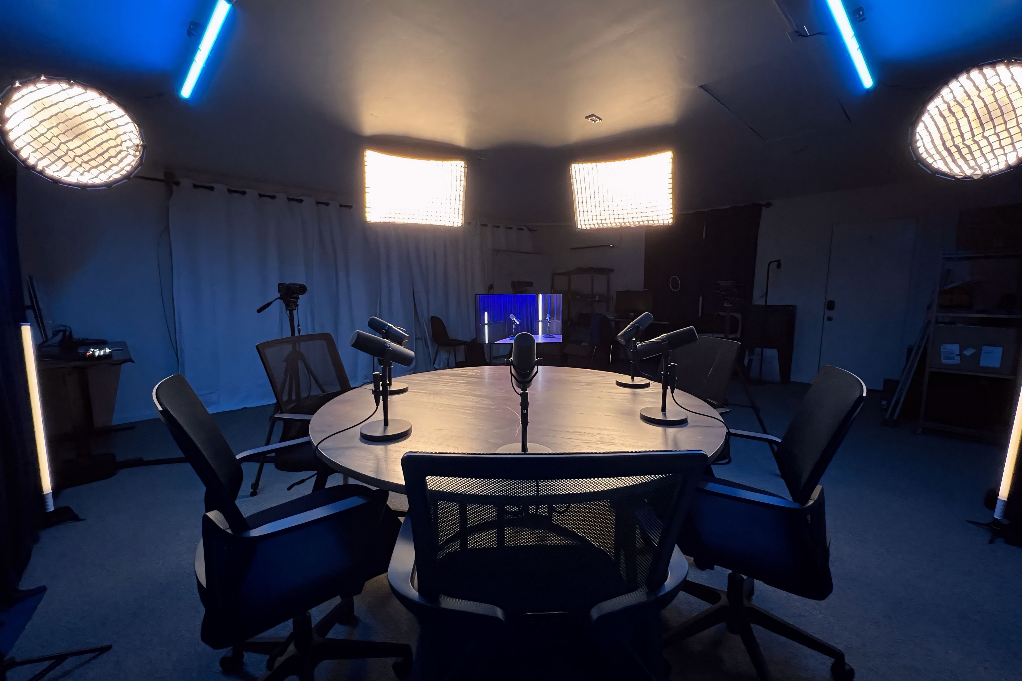 A wide-angle shot of a professional podcast studio centered around a large, round wooden table. Five Shure SM7B microphones on desk stands are arranged at seating positions, accompanied by five black mesh office chairs. The lighting setup features four warm Aputure softboxes: two rectangular panels hanging directly above the table and two circular grids suspended on the far left and right. Blue LED tube lights are mounted on the ceiling and placed vertically on the floor to provide cool accent lighting. In the background, white curtains, a camera on a tripod, a production monitor, and equipment shelves are visible in the dimly lit room.