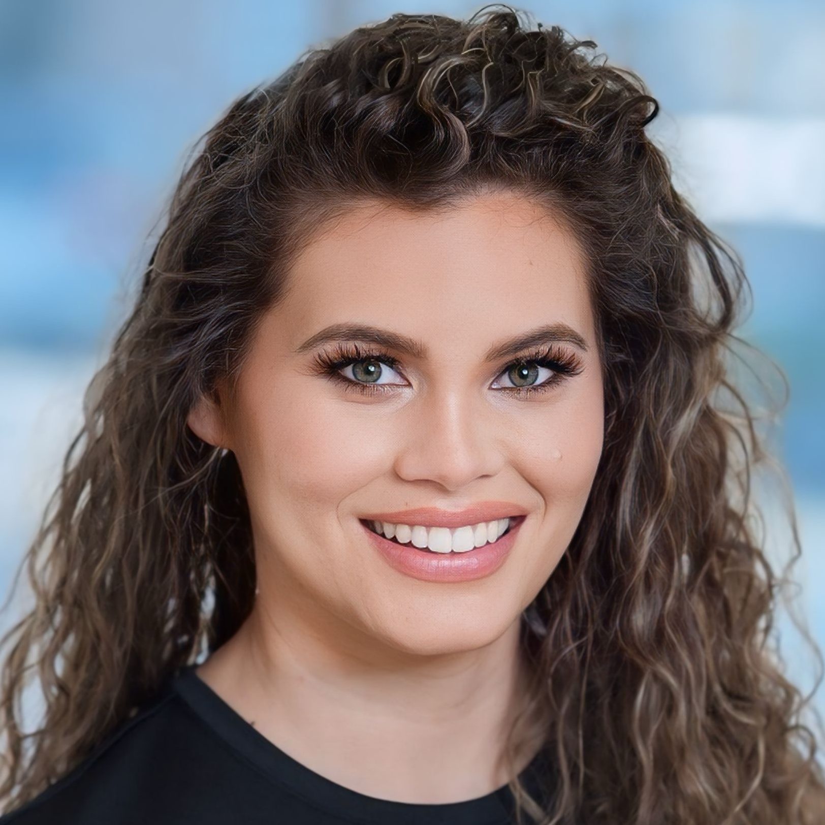 A headshot of a woman with curly brown hair and green eyes, smiling broadly at the camera. She wears dark mascara and a black top, against a blurred blue and grey background.